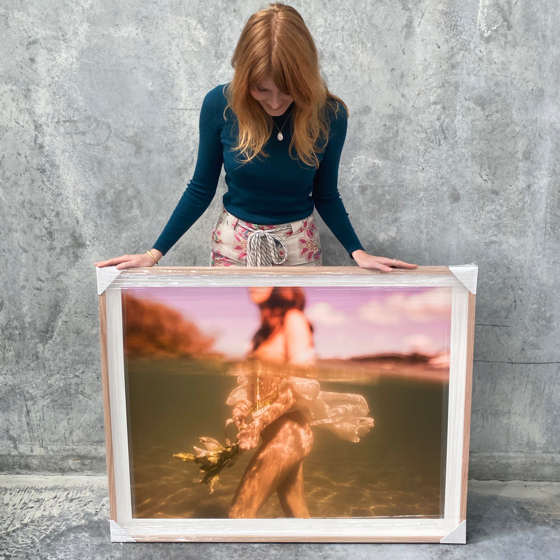 A woman with long red hair, dressed in a dark green top and floral skirt, stands against a concrete wall holding a large framed photograph. The photograph, titled "The Language of Flowers #1" by Francesca Owen, is printed with archival pigments on fine art paper and depicts an underwater scene with a blurred figure holding flowers.