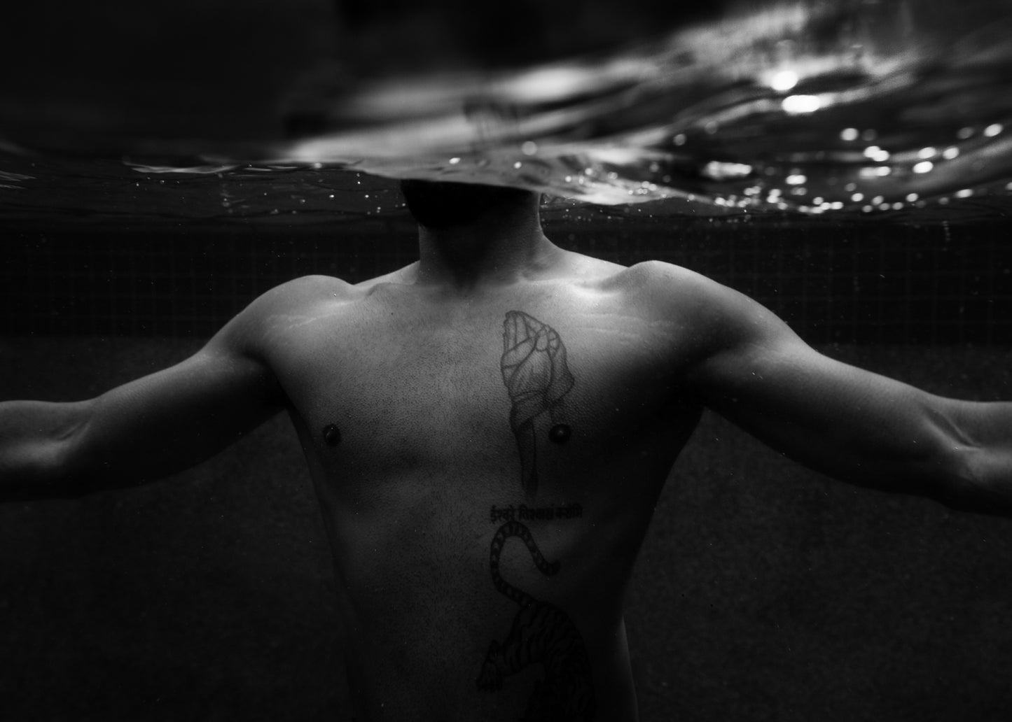 The captivating image titled "New Waters #1" by Francesca Owen features a black and white photograph of a man's torso submerged underwater, with his head above the water's surface. The man has outstretched arms and visible tattoos on his chest and lower abdomen, including a raised fist, text, and a snake design. This artwork is available on fine art paper with worldwide shipping.