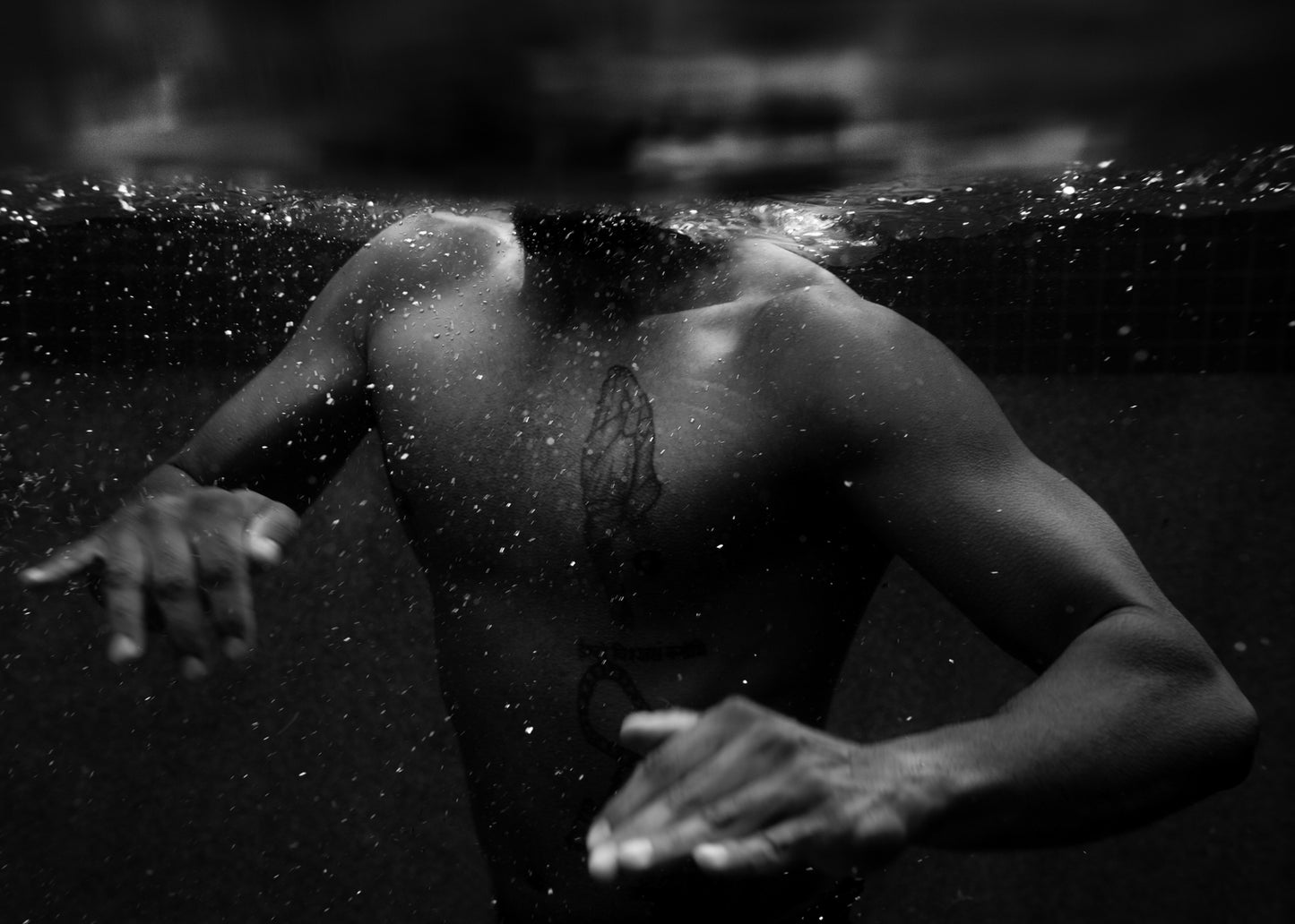 New Waters #2 by Francesca Owen features a black-and-white photo of a person submerged in water up to their neck, with only their chest and tattooed arms visible. Gentle surface ripples are surrounded by tiny air bubbles. Captured on fine art paper using archival pigments and hand-signed, this timeless work of art is a stunning addition to any collection.