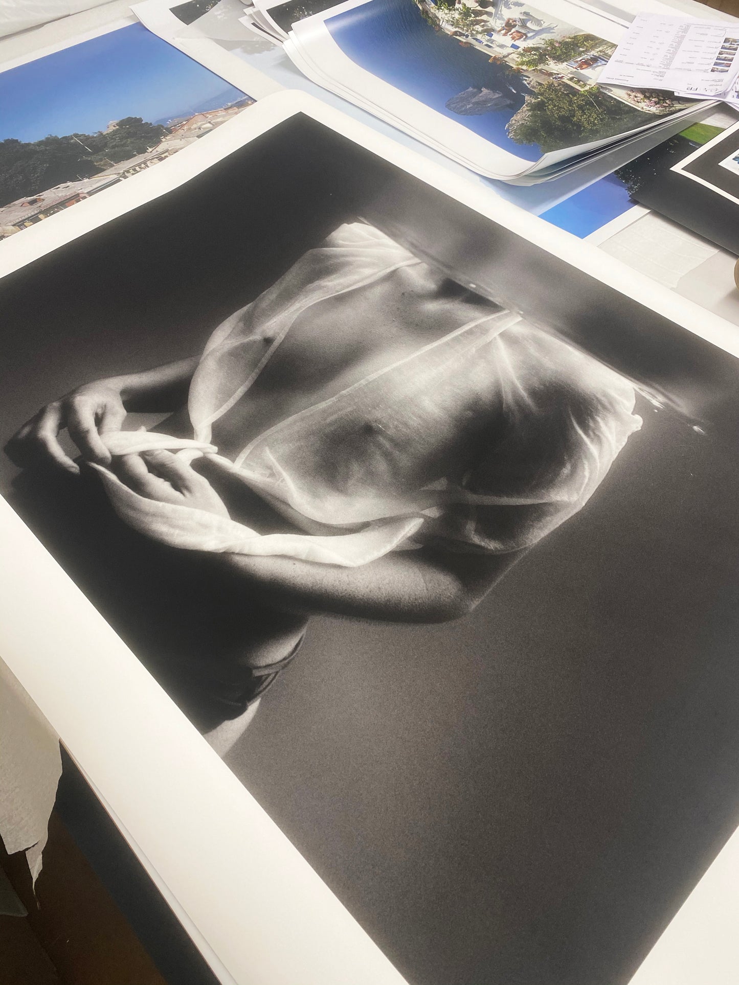 Close-up photo of "Softness #2" by Francesca Owen, an artistic black-and-white image featuring a person viewed from the shoulders down, wearing a sheer top. This striking image, printed with archival pigments on fine art paper, is laid out on a table alongside various other photographs and printed materials in the background.