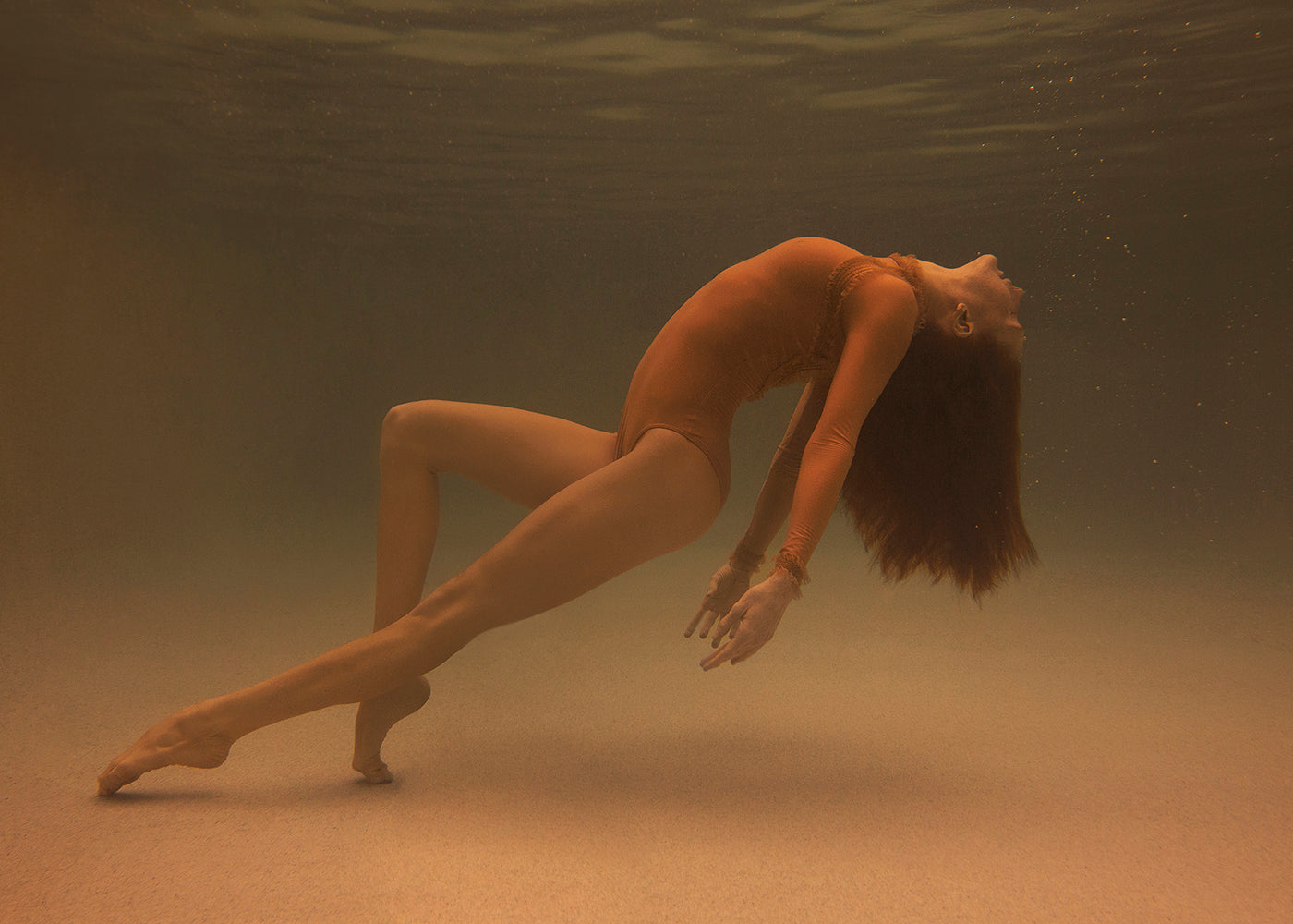 Beneath the water's surface, a figure from Francesca Owen's Open Hearted collection elegantly arches their back in an orange bodysuit. With their head tilted upwards, legs extended, and toes pointed, they create a dynamic and graceful pose against a softly illuminated background. Bubbles trail from their lips as they move.