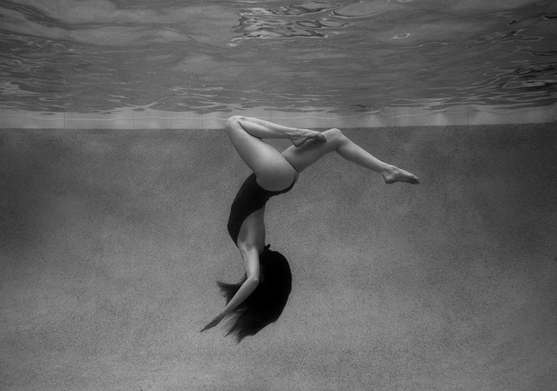 A black-and-white underwater photo, Palm Springs Solo by Francesca Owen, captures a person in a swimsuit performing a backward somersault. Suspended upside down with bent knees and arms extended, their long hair flows downward as the rippling surface of the water is visible above. Available in custom sizes and framing options, this print uses archival pigments for lasting quality.