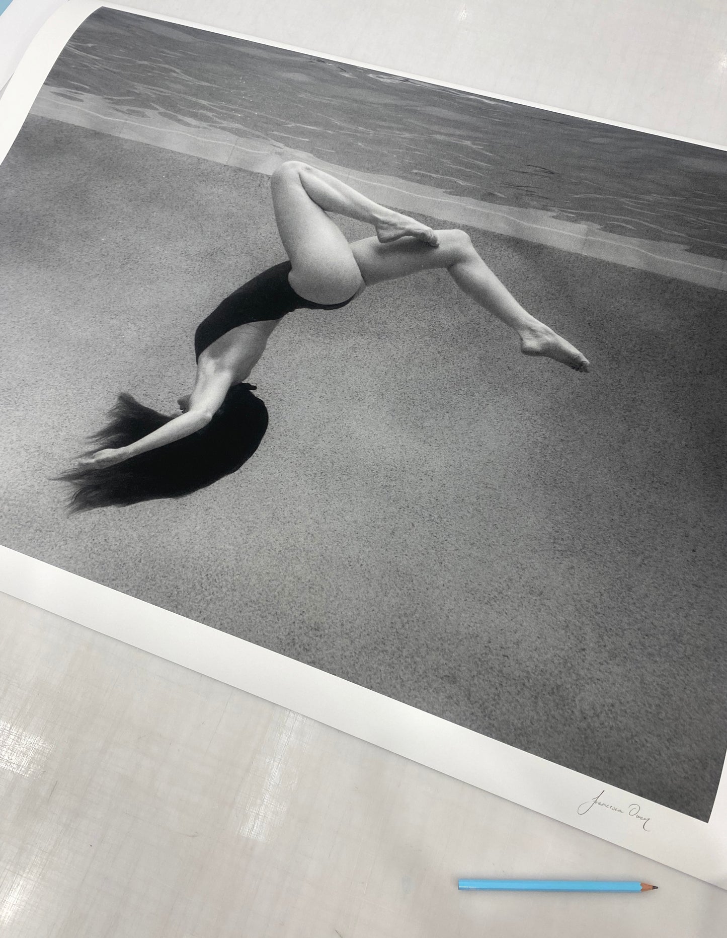 In "Palm Springs Solo" by Francesca Owen, a black and white photograph printed on fine art paper captures a person mid-backflip in a swimming pool, their long hair flowing and body curled. The image is signed by the artist in the bottom right corner, while a blue pencil rests nearby on the surface.