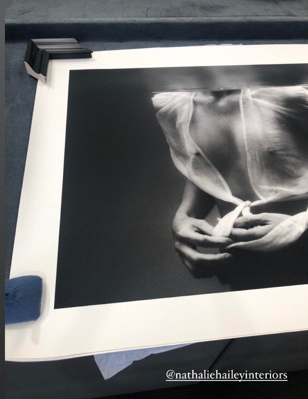 The artwork titled "Softness #2" by Francesca Owen features a black-and-white photograph on fine art paper, depicting a partially open sheer fabric that reveals shoulders and a chest. Accompanied by a certificate of authenticity, the print is displayed flat with black frame corners at the top. A blue cloth lies beside it, with the text "@nathaliehaileyinteriors" visible at the bottom.