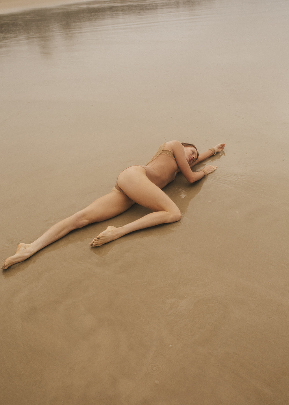 A person is lying face down on the wet sand of a beach, with their arms bent and their left leg slightly bent. They are wearing a bikini bottom and appear relaxed, with their eyes closed. The serene beach scene captures an almost ethereal quality, reminiscent of Francesca Owen's fine art prints from the Red Head Series #3.