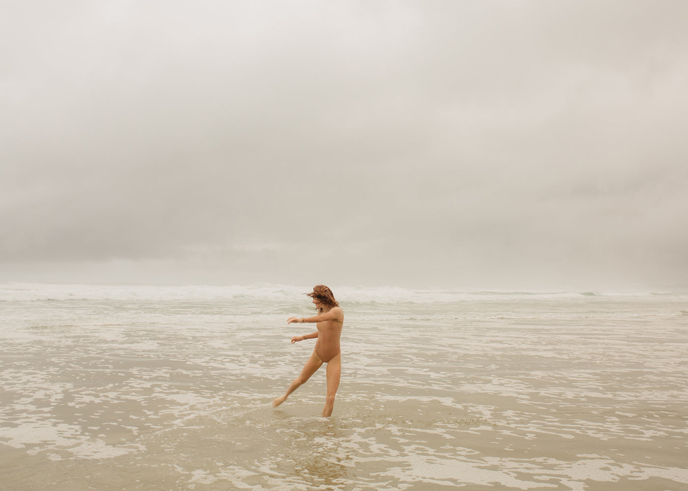 A serene and natural scene is captured in *Red Head Series #5* by Francesca Owen, depicting a nude individual standing in shallow waters on a beach. The person, with arms outstretched, faces slightly to the left amidst gentle waves under a cloudy, overcast sky. This artwork utilizes archival pigments on fine art paper to bring the moment to life.