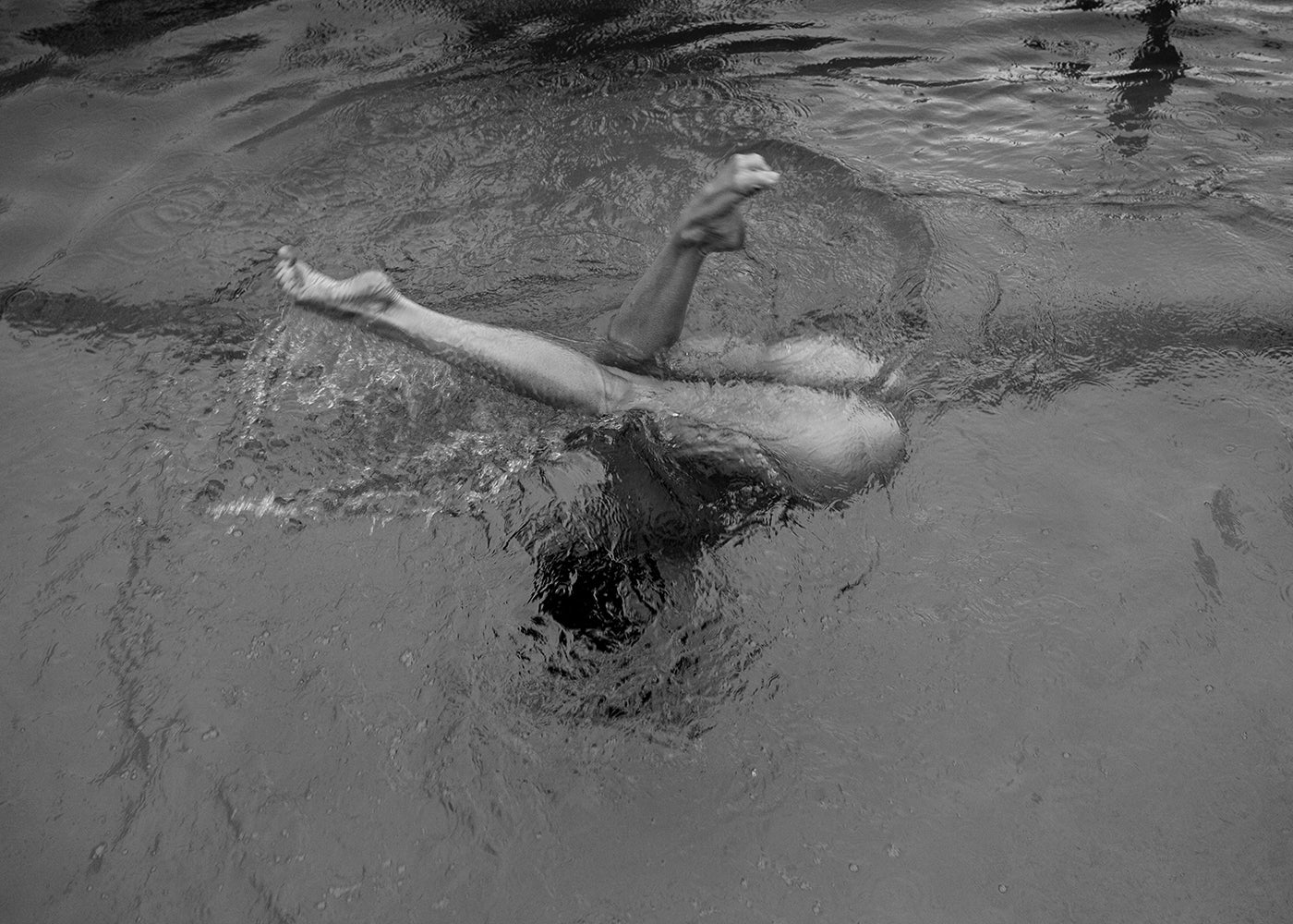 Description: "Red Head Series #8" by Francesca Owen features a black and white photo of a person swimming underwater, with only their legs visible above the surface, creating a splash. The surrounding water ripples indicate movement, capturing the dynamic motion of the swimmer. This scene is printed with archival pigments on fine art paper for discerning collectors.