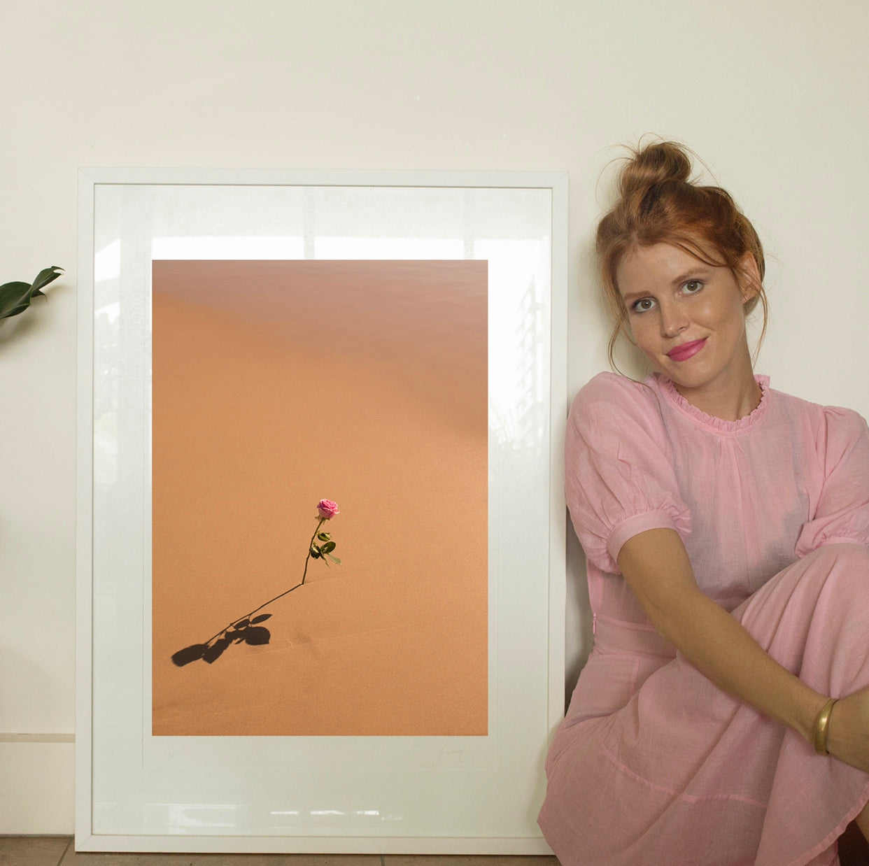 A red-haired woman in a pink dress sits on the floor next to a framed photo titled "Portrait of a Rose" by Francesca Owen, depicting a single pink rose casting a shadow on a sandy desert background. The white wall behind them sets off a minimalistic and serene scene. This artwork is available as limited edition prints on fine art paper with worldwide shipping.