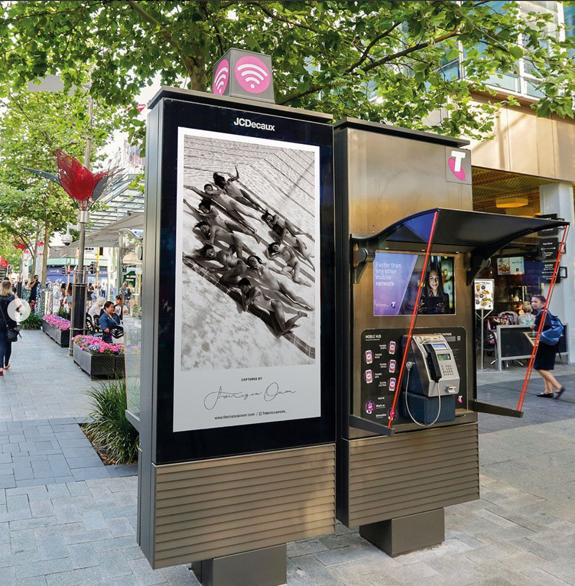 On a bustling city street, a public phone booth prominently displays a large advertisement poster for a ballet performance. Pedestrians stroll by on the sidewalk, while planters brimming with flowers and shade-giving trees add to the charm of the scene. Captured by fine art photographer Francesca Owen, this captivating moment is available as a limited edition print titled "Ladies in Waiting #2" on giclee fine art paper.