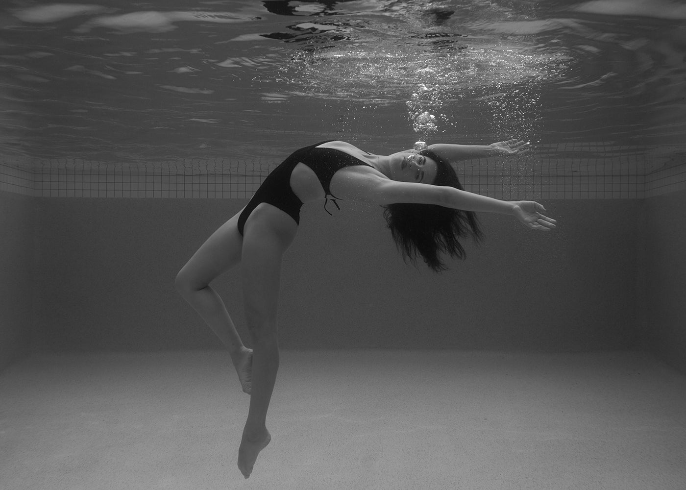 Self Reflection 02 by Francesca Owen captures a black and white image of a woman in a one-piece swimsuit swimming underwater. She gracefully arches her back with her arms extended, bubbles rising from her mouth. The background reveals the tiles and smooth floor of the pool. This piece is printed on fine art paper with archival pigments and comes with a certificate of authenticity.