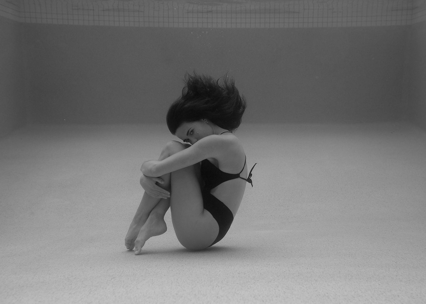A woman in a black swimsuit is underwater in a pool, curling up into a tight ball with her arms wrapped around her legs. Her dark hair floats freely in the water, and the pool floor is visible beneath her. This black-and-white image, titled "Self Reflection 12" by Francesca Owen, is printed on fine art paper with archival pigments and comes with a certificate of authenticity.