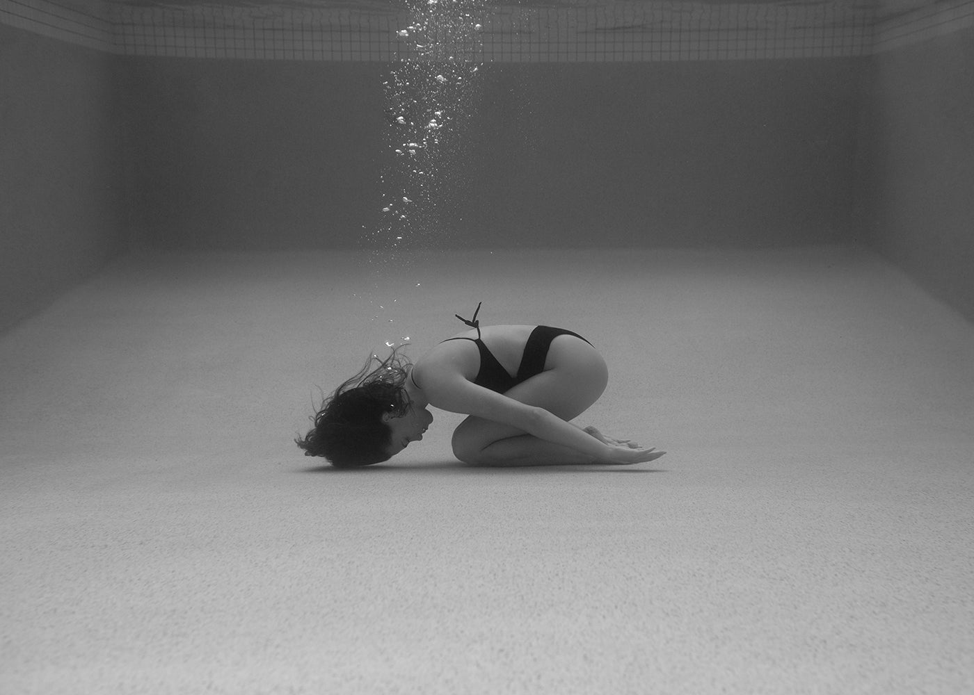 A person in a black swimsuit curls up on an empty pool’s floor underwater, bubbles rising above—echoing the striking composition of Francesca Owen’s hand-signed art print, Self Reflection #16.