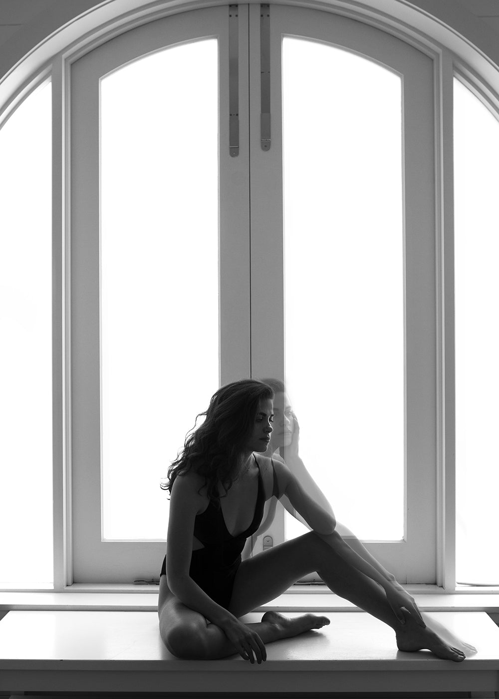 A woman in a black bodysuit poses thoughtfully on a windowsill before a bright arched window. "Self Reflection #9" by Francesca Owen is available as hand-signed fine art prints on paper.