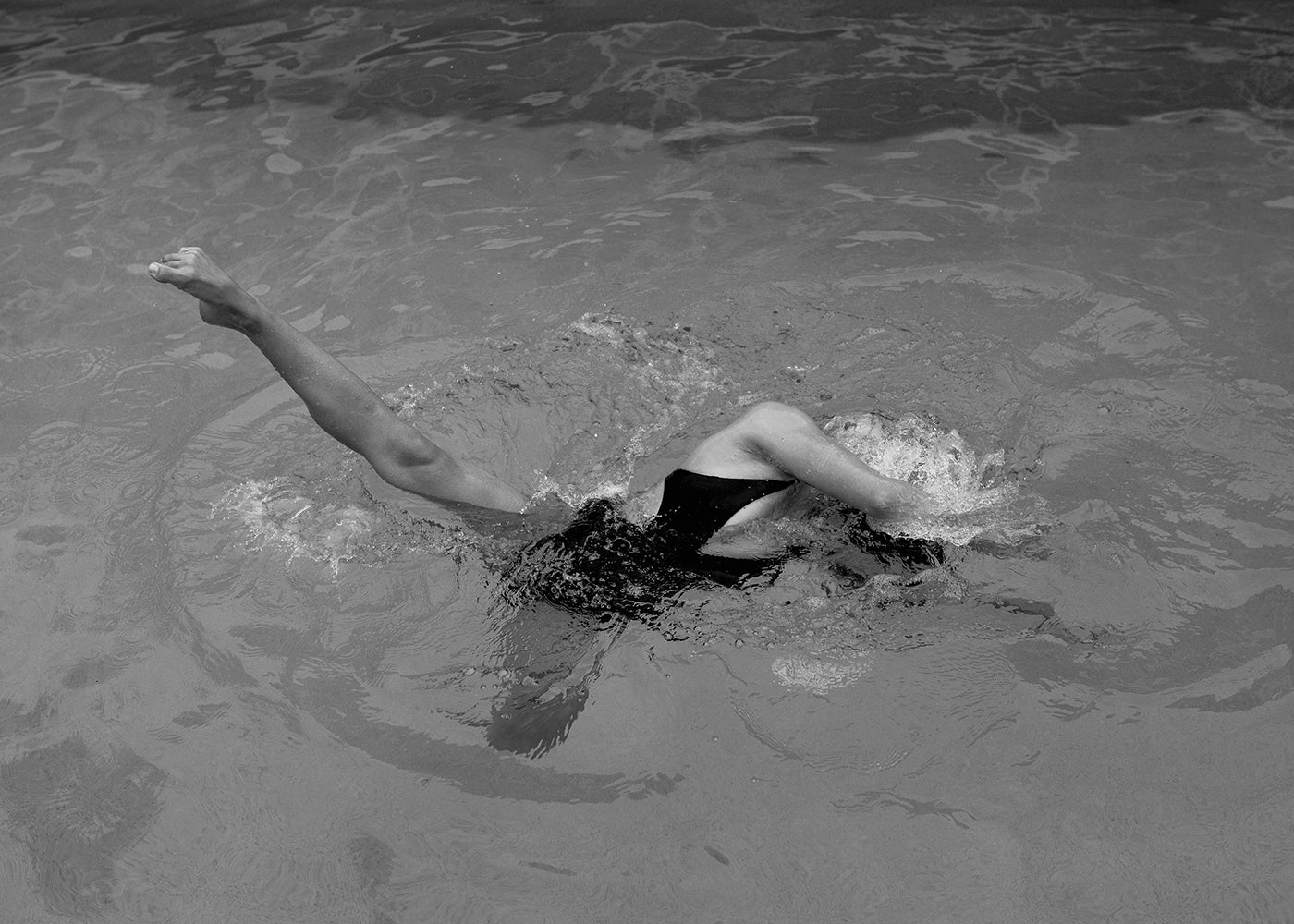 A striking piece titled "Self Reflection 18" by Francesca Owen, featuring a black and white image of a person swimming on their back in a body of water. One leg is extended straight up while the other is bent at the knee. Their arms are spread out, creating ripples on the water’s surface around them. This evocative scene is elegantly rendered with archival pigments on fine art paper.