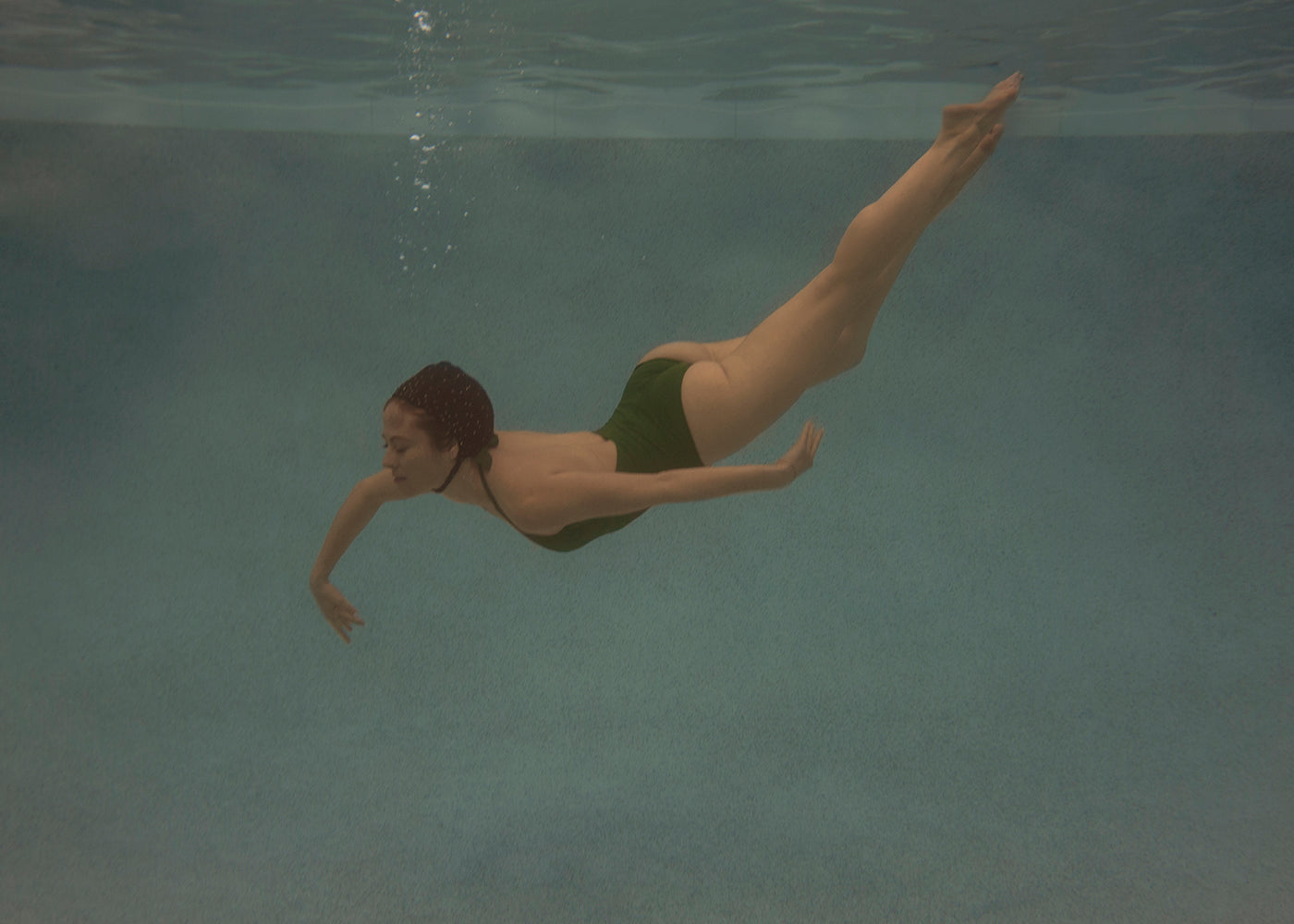 A person in a green swimsuit is swimming underwater in a pool. Their streamlined body and extended limbs create a graceful pose, with bubbles trailing from their mouth as they hold their breath. The pool's blue water provides a serene backdrop, reminiscent of Francesca Owen's fine art print "Silent Surrender #2.