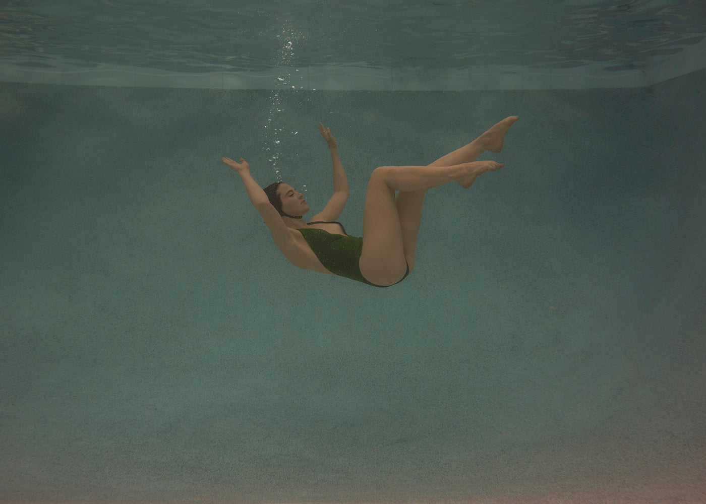A person in a black swimsuit is submerged underwater, facing upwards with arms and legs bent and raised. Bubbles rise from their mouth and nose as the light gently illuminates the surrounding blue water. The serene scene captures a moment of tranquility, reminiscent of "Silent Surrender #1" by Francesca Owen, as if printed on fine art paper with archival pigments.