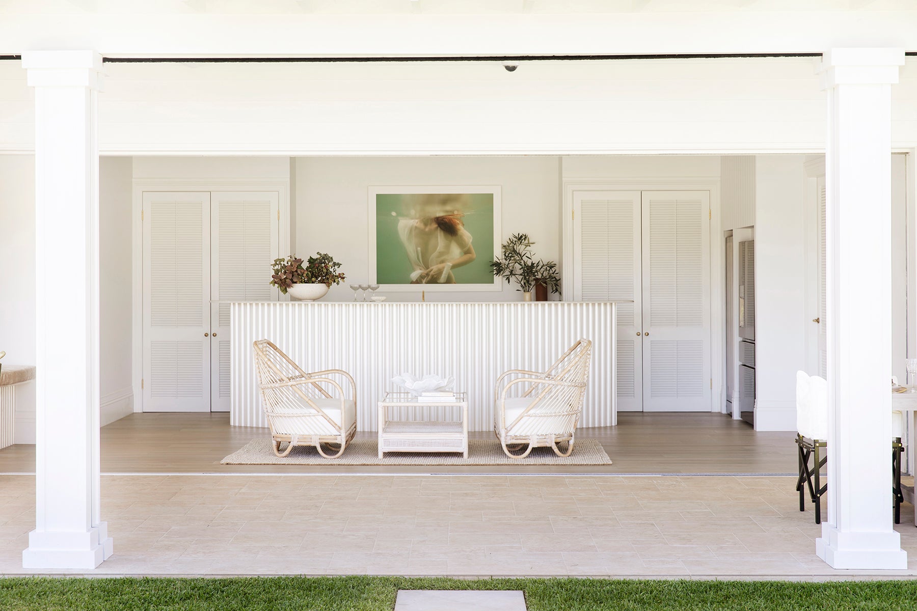 A bright, airy indoor space with white décor. Two white wicker chairs face a white table, positioned in front of a white-paneled wall. The wall features "Softness #1" by Francesca Owen, a large photo of a swimmer printed with archival pigments on fine art paper. Plants and decorative items are placed on a shelf above the panels.