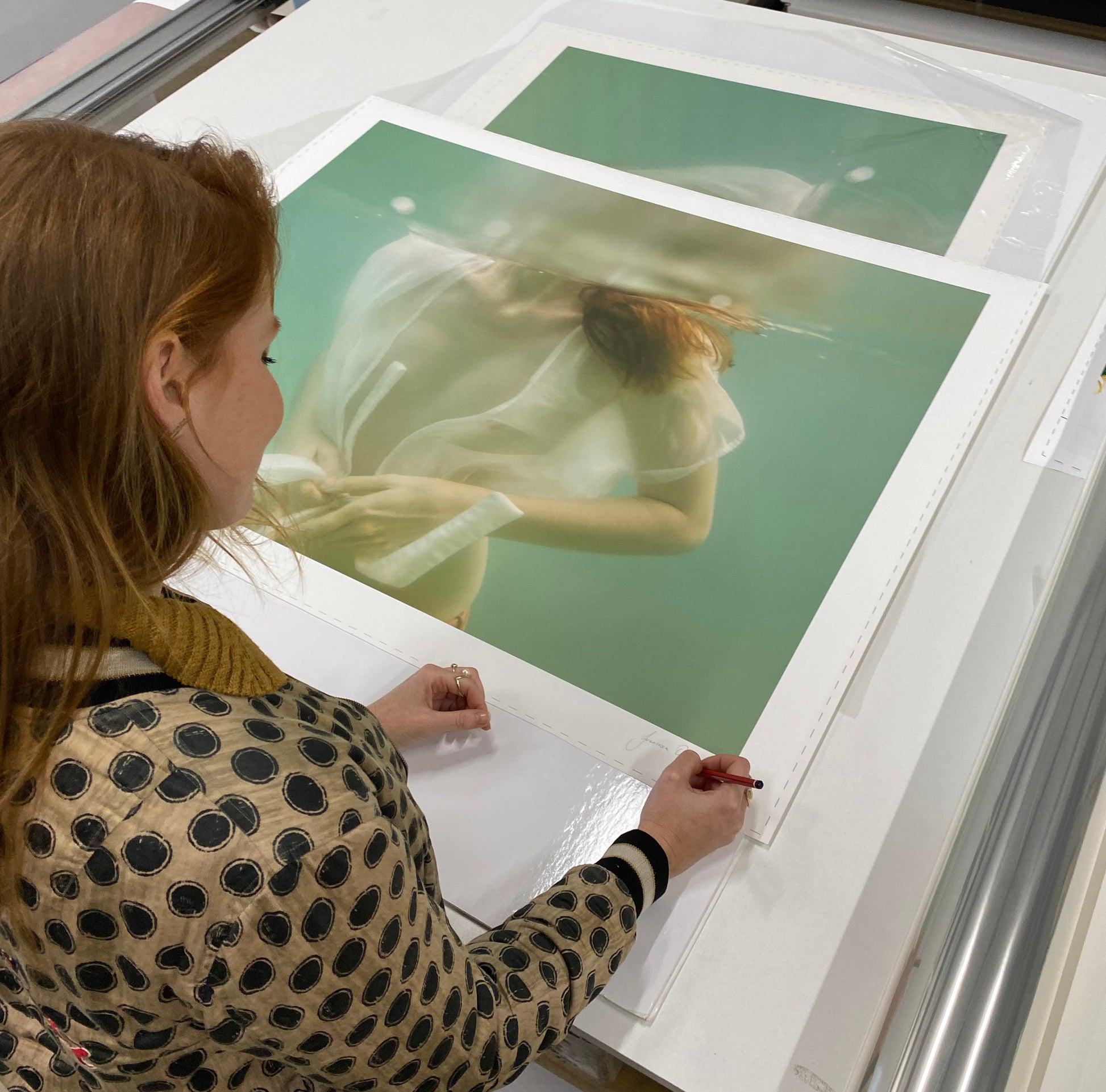 A person with reddish-brown hair, wearing a polka-dotted sweater, is carefully examining and possibly signing a large photograph of an underwater scene titled "Softness #1" by Francesca Owen, printed on fine art paper. The person is leaning over a table covered in art prints, ensuring the image retains its vibrancy through archival pigments.