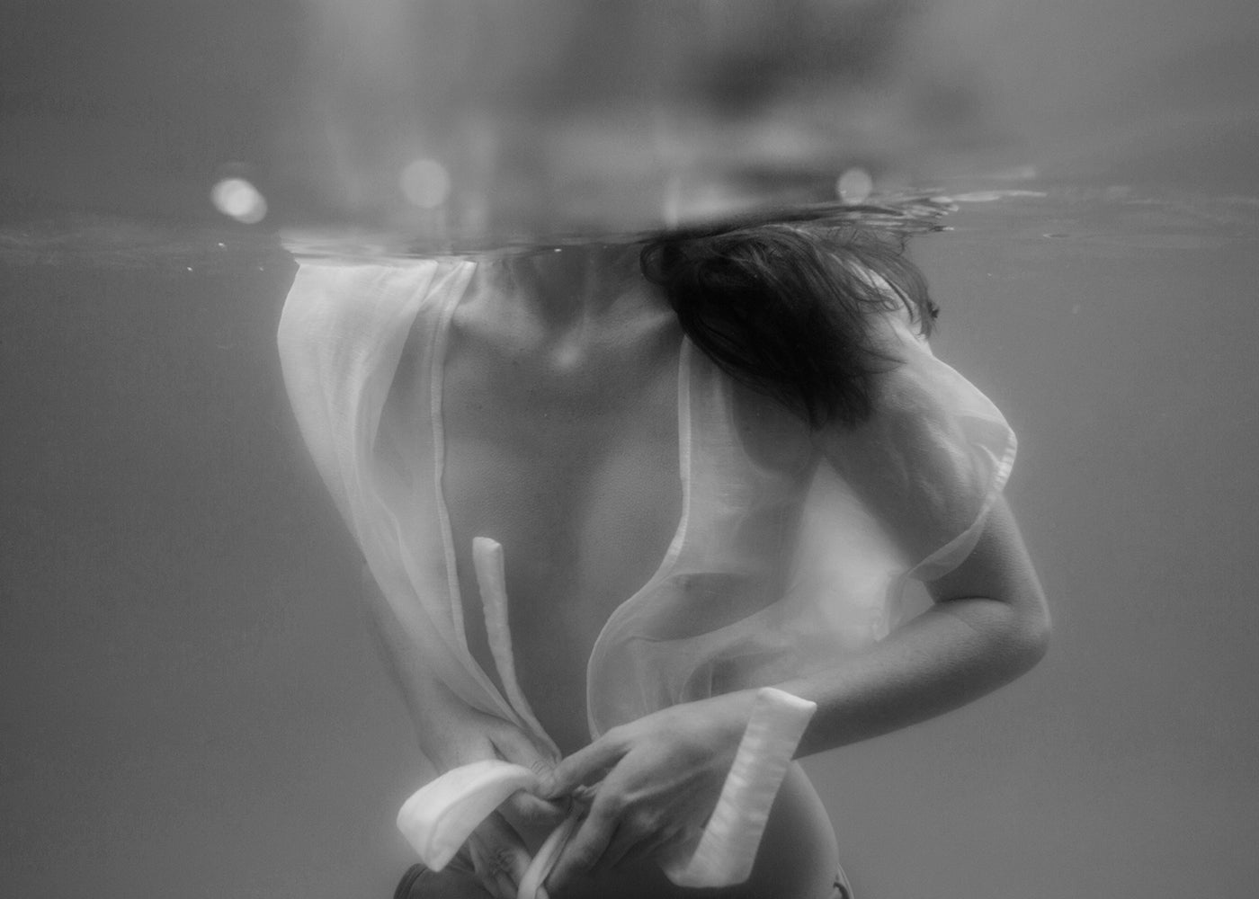 The intriguing black and white underwater photograph, titled "Softness #1 B&W" by Francesca Owen, captures a person partially submerged in water wearing an unbuttoned, translucent white shirt. With their head above the water's surface and their torso and hands below, the image creates an ethereal and serene atmosphere ideal for fine art prints or custom framing.