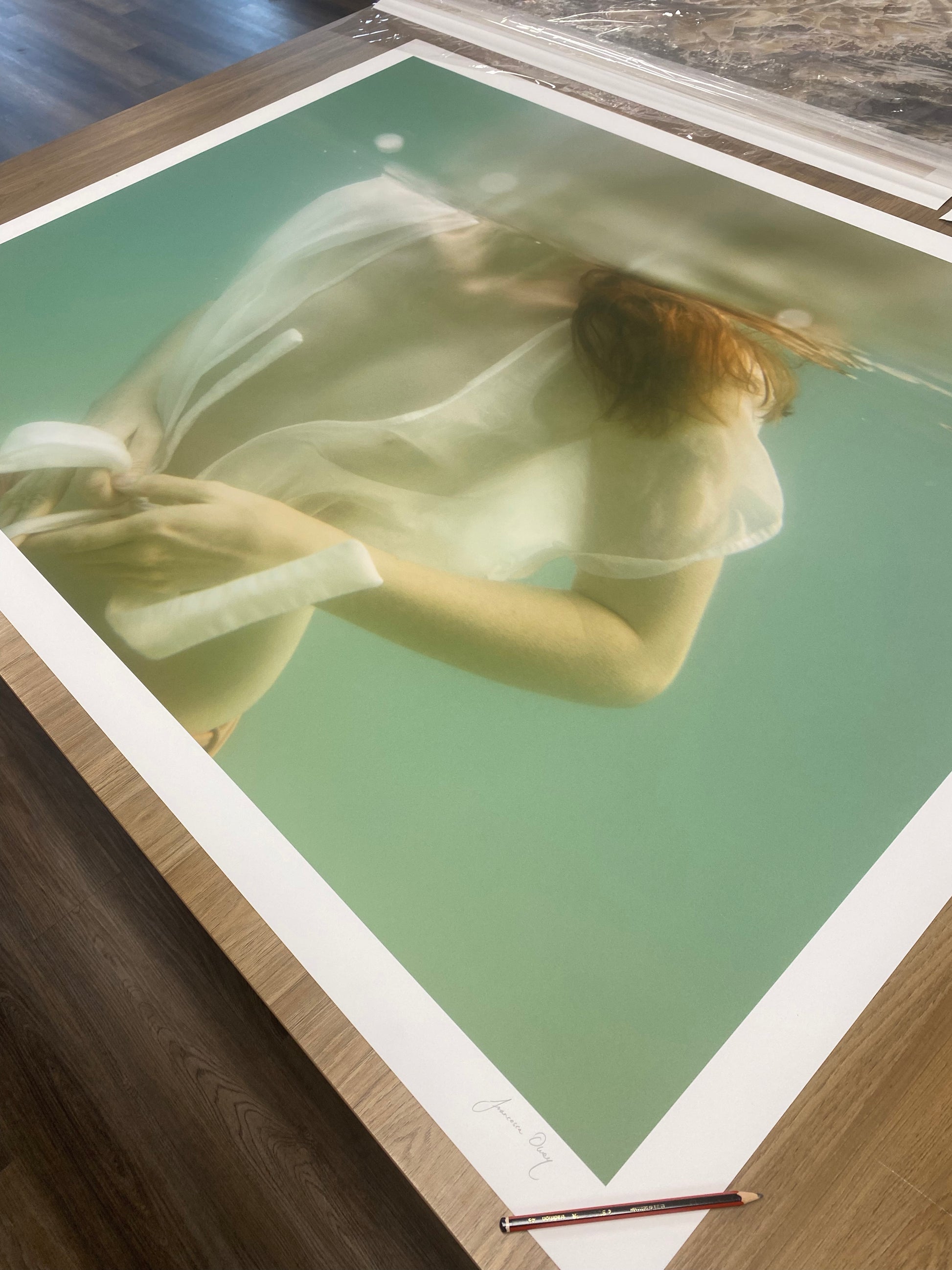 A large photographic print titled "Softness #1" by Francesca Owen shows a person underwater, partially obscured by the water's surface. The individual wears a sheer, white, flowing garment. A pencil rests on the table near the edge of the print, which has an indistinct signature and comes with a certificate of authenticity.