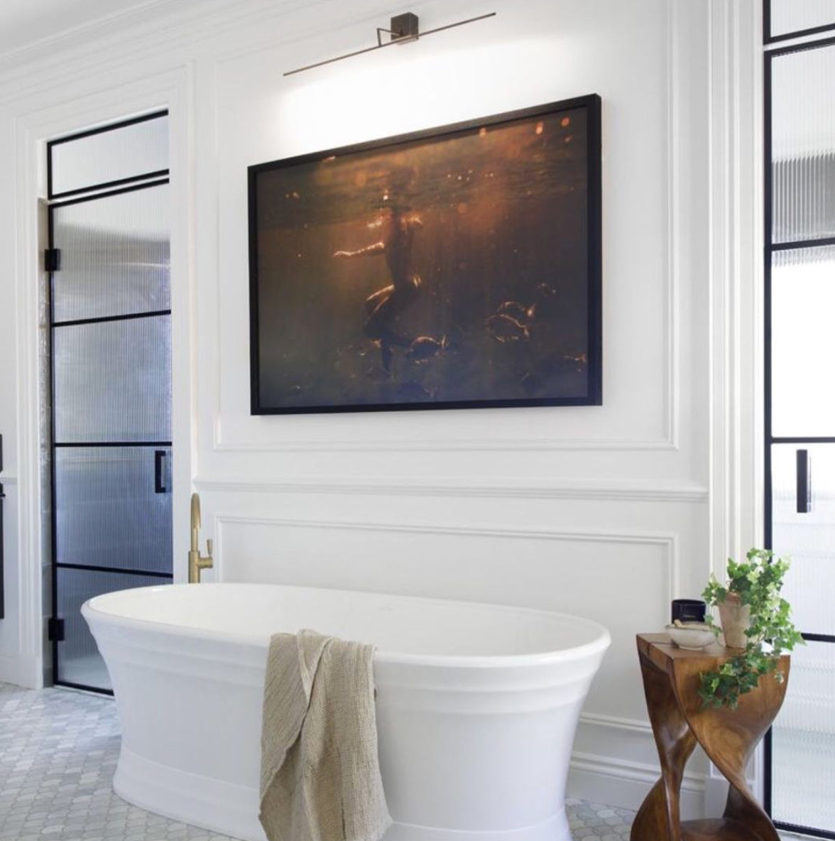 A modern bathroom showcases a white freestanding tub adorned with a beige towel draped over its edge. Above the tub hangs "Still Waters #2" by Francesca Owen, a large framed painting of a person underwater, rendered on art paper using archival pigments. A small wooden side table with a potted plant is positioned next to the tub, while frosted glass doors provide a serene backdrop.