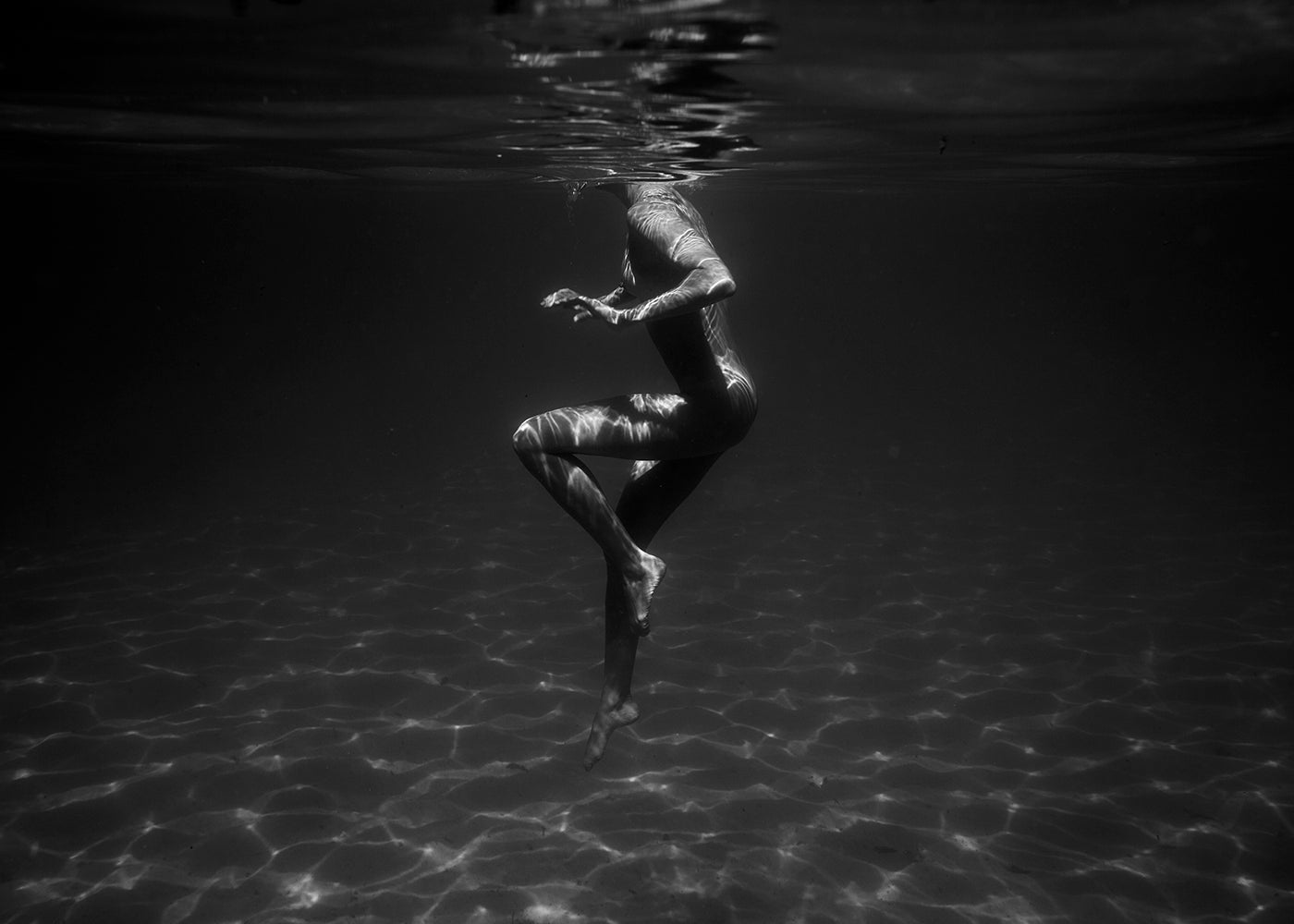 Still Waters #1 by Francesca Owen features a mesmerizing black and white underwater photograph of a person submerged in water, bathed in light filtering from above which creates ripples on their body. The figure is depicted in a dynamic pose, partially upside down with bent legs and extended arms. This stunning piece is printed on fine art paper using archival pigments and is available for worldwide shipping.