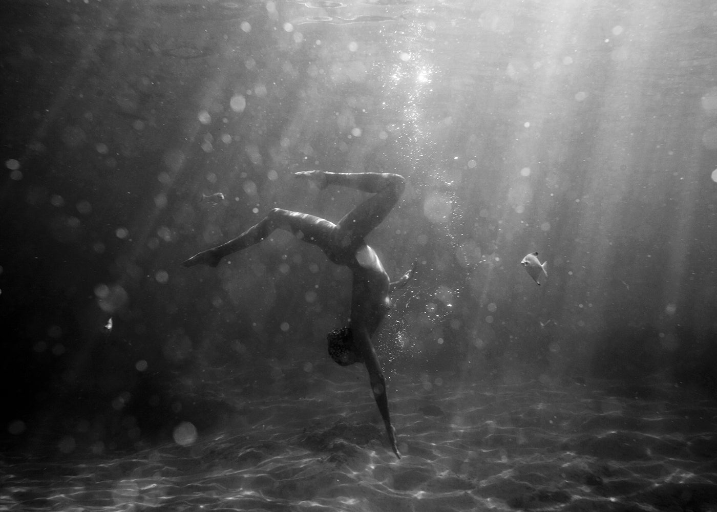 A person performs an underwater handstand amid bubbles and sunlight rays, creating a dreamy scene in "Still Waters #3," a fine art paper print by Francesca Owen.