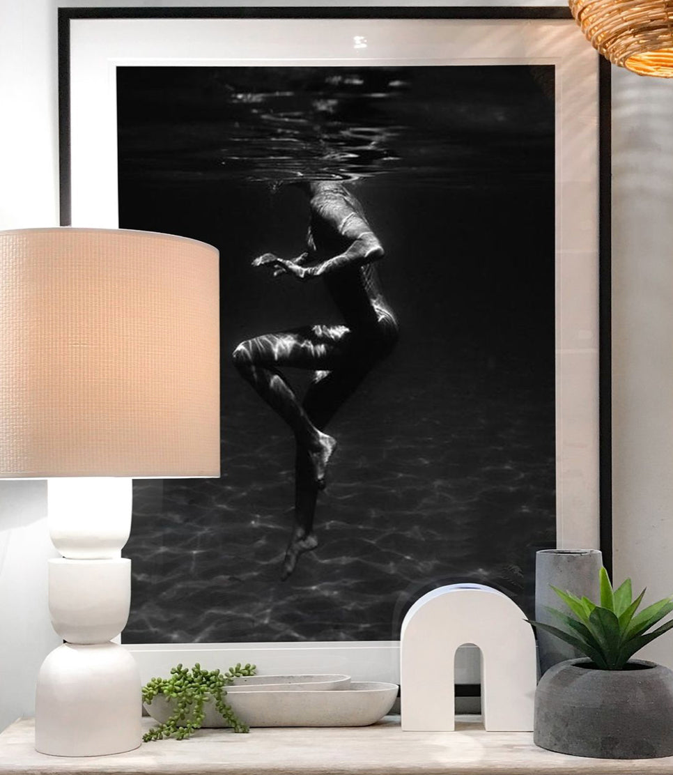 A modern, well-lit room features "Still Waters #1," a monochrome photograph by Francesca Owen depicting a person swimming underwater, printed on fine art paper with archival pigments. The room includes a round white table lamp, minimalistic decor such as small plants, and a geometric white sculpture. The overall aesthetic is clean and contemporary.