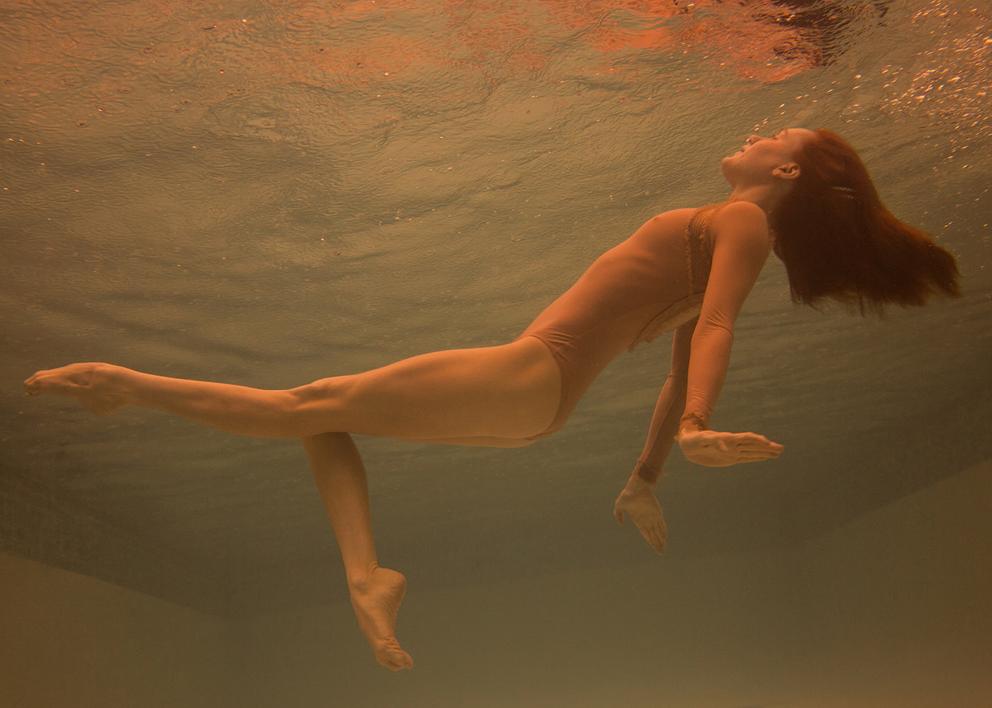 A woman in the "Surfacing" one-piece swimsuit by Francesca Owen is underwater in a pool, her eyes closed and arms outstretched. Her hair and body are gracefully floating, conveying a serene sense of swimming or floating. The warm, sepia-toned lighting creates an ambiance perfect for capturing as Limited Edition fine art prints.