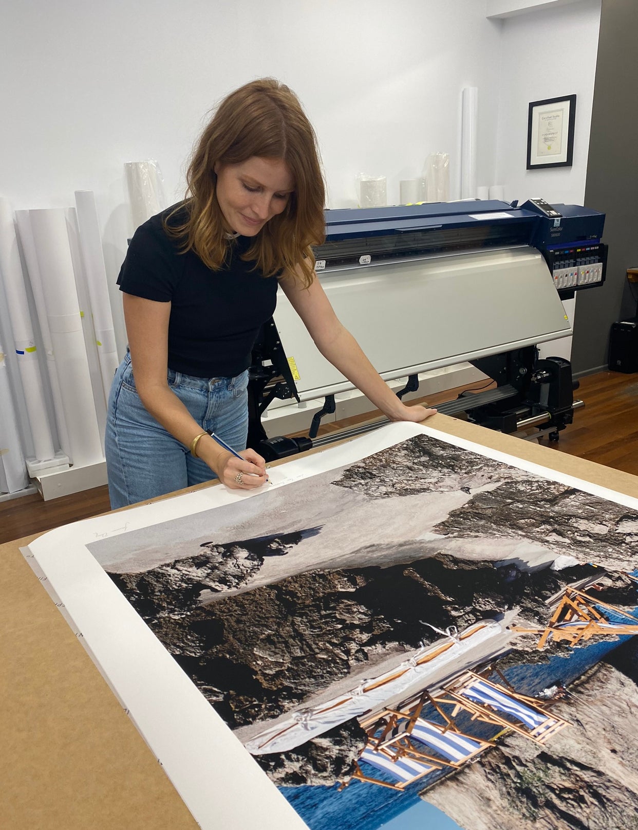 A woman with shoulder-length brown hair wearing a black shirt and blue jeans is signing "Waiting For You #1" by Francesca Owen, a large coastal scene photograph featuring rocks and ocean water, which is printed using archival pigments on fine art paper. She is in a room with a large printer and rolls of paper in the background.