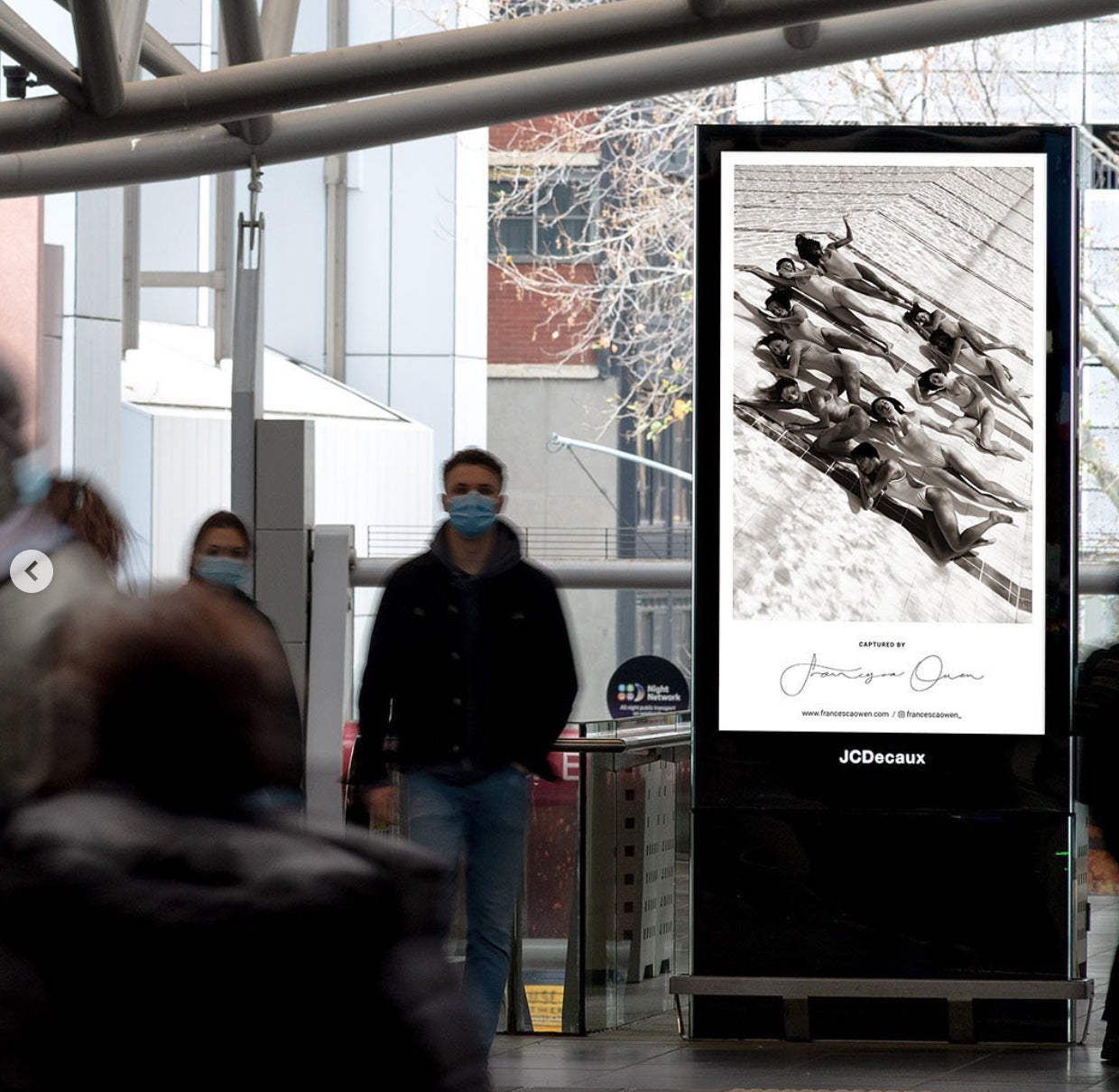 A digital billboard inside a modern building displays "Ladies in Waiting #2," a black-and-white image of synchronized swimmers by the renowned fine art photographer Francesca Owen. Several people, some wearing face masks, walk by the billboard. The building has large windows and metal beams, adding to its contemporary ambiance.