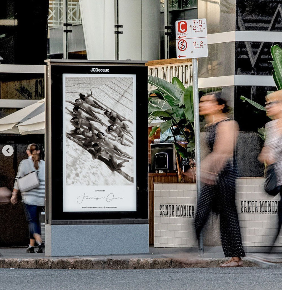 A sidewalk scene with people walking past an advertising display case featuring a black and white photograph titled "Ladies in Waiting #2" by fine art photographer Francesca Owen. The case, displaying a limited edition print on giclee fine art paper, is situated near a building with "Santa Monica" signage visible in the background.