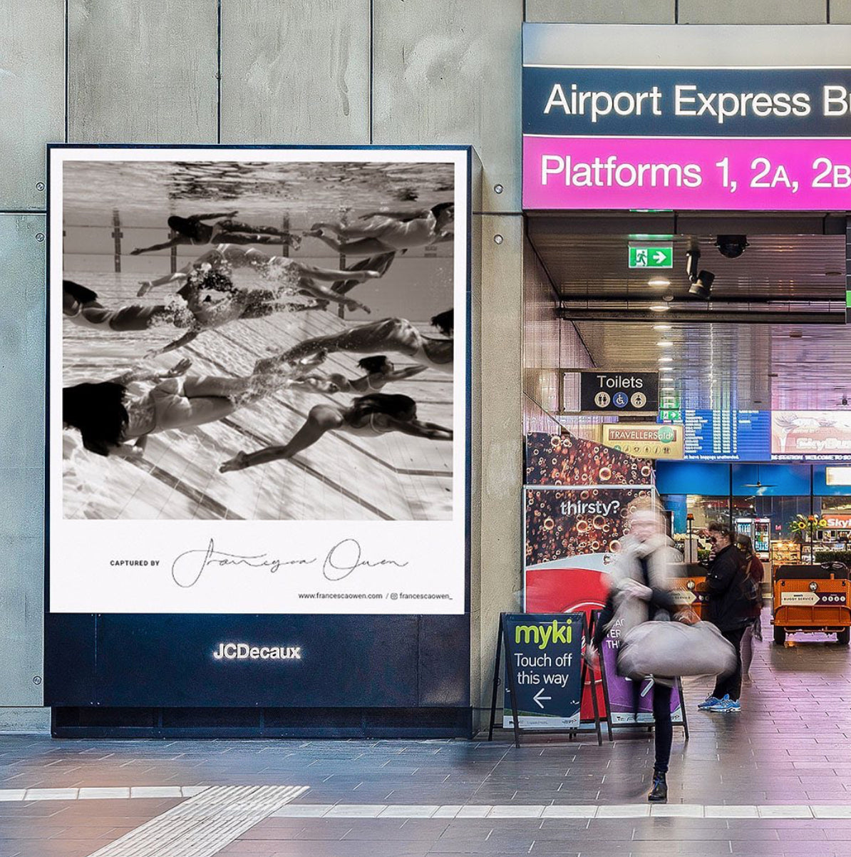 A busy train station features a digital billboard displaying "Ladies in Waiting #4," an underwater black-and-white photograph by the fine art photographer Francesca Owen. The station has signs for "Airport Express" and platforms 1, 2A, and 2B. A person carrying a bag walks past the limited edition billboard towards platform signs.
