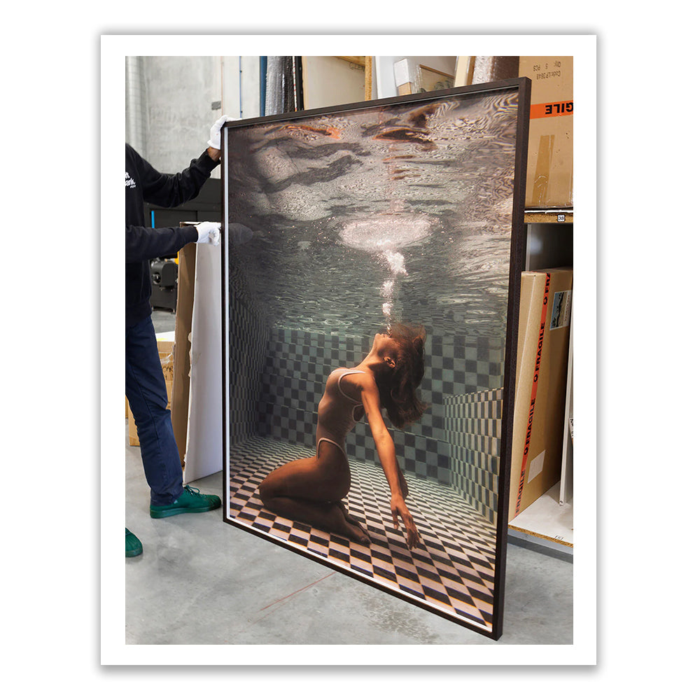 A woman in a white swimsuit kneels and leans back underwater in a tiled pool, exhaling bubbles towards the surface.