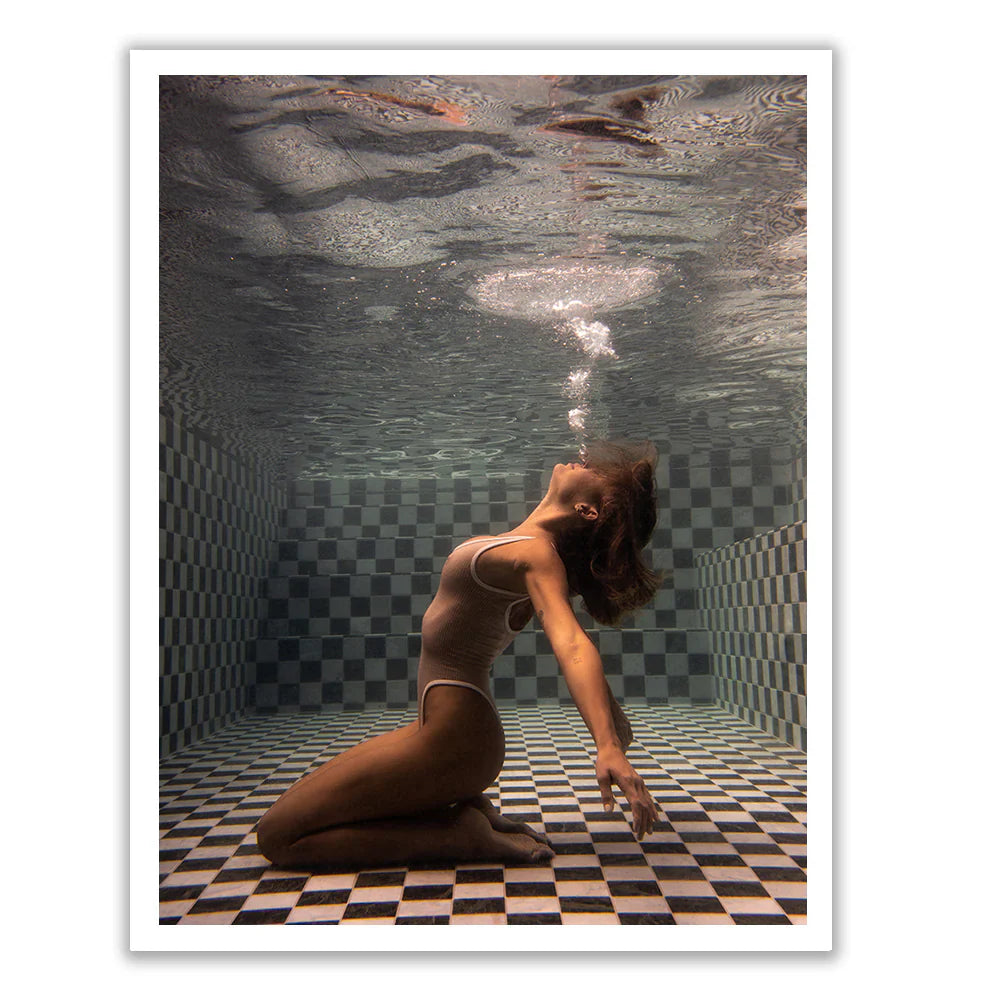 A woman in a white swimsuit kneels and leans back underwater in a tiled pool, exhaling bubbles towards the surface.