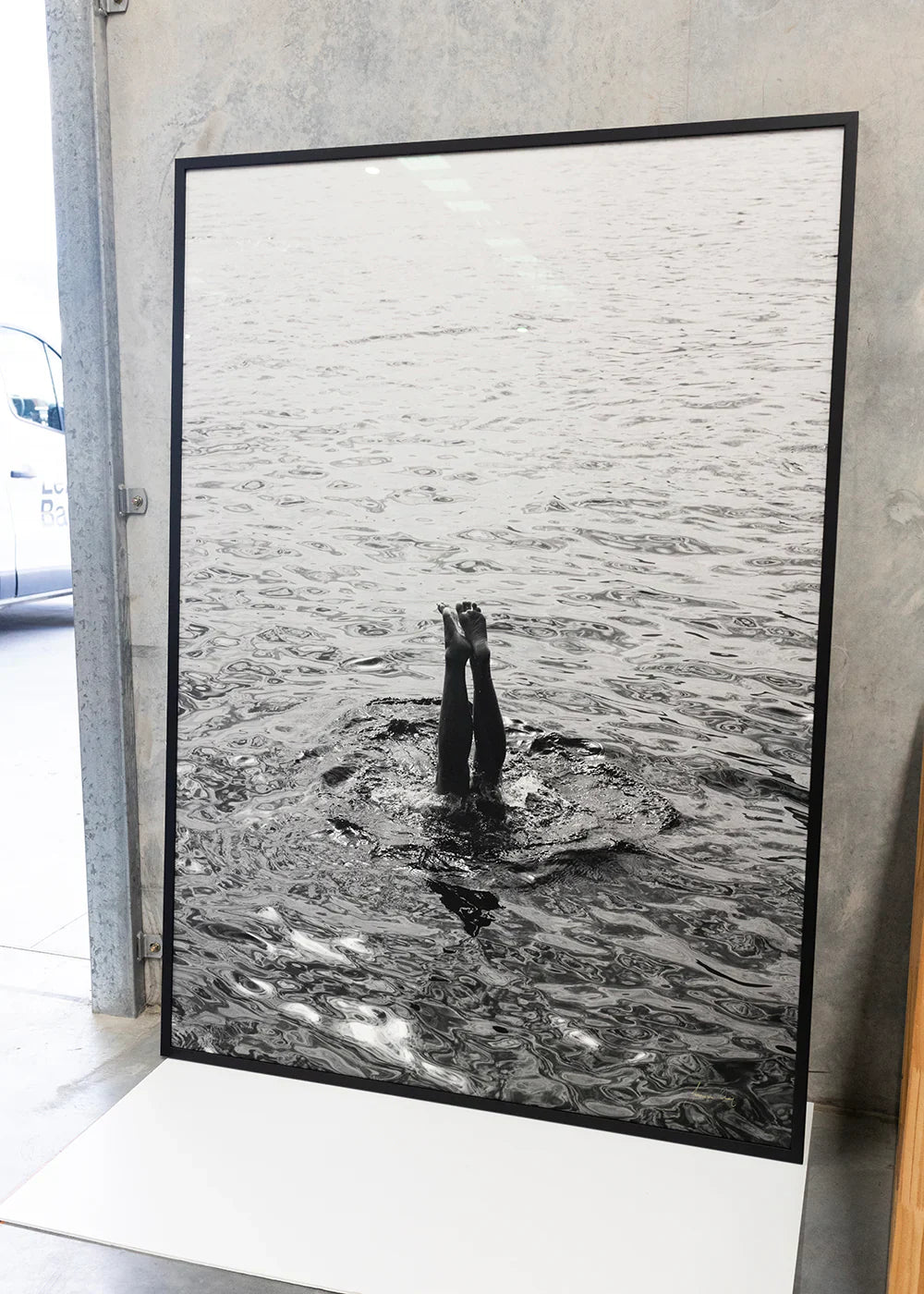 A minimalist bedroom features a white bed, gray headboard, and a side table with a lamp. A large framed black-and-white photo hangs on the wall. An open door leads to a bathroom with tiled walls.