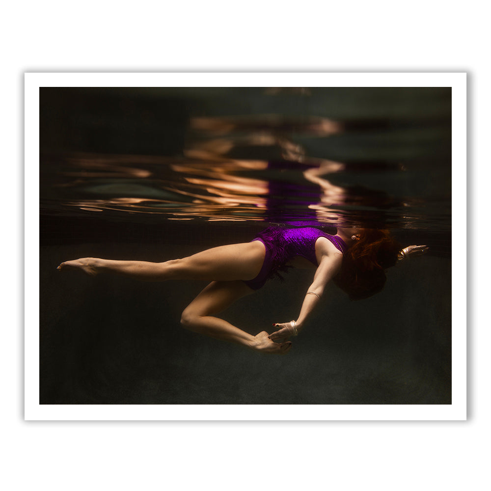 A person in a purple swimsuit is floating underwater, mirroring their reflection on the surface above. The scene has a serene, artistic quality with warm lighting.