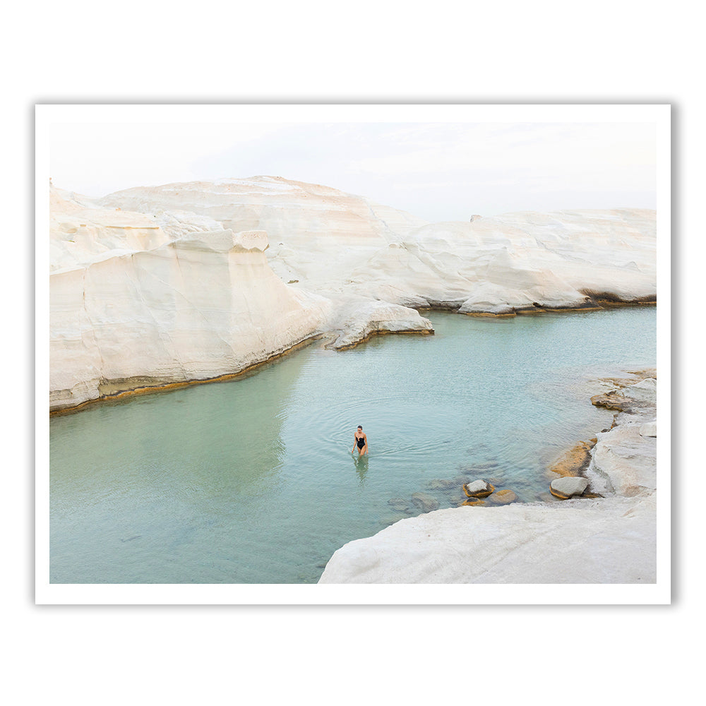 A person stands in the calm, clear waters of a natural pool surrounded by smooth, white rock formations, under a pale sky.