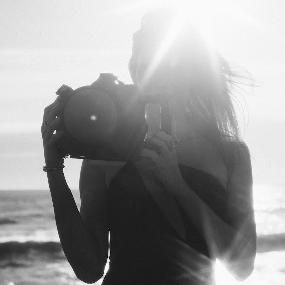 A woman in a strapless swimsuit holds a large underwater camera housing, standing in shallow water with a rocky background. The photo is in black and white.