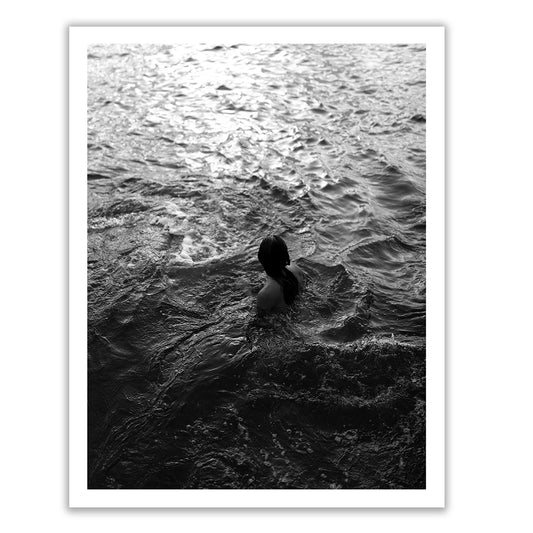 Sunrise Swim #11 by Francesca Owen captures a black and white photo of a person in the ocean viewed from behind. The individual stands waist-deep in the water, surrounded by rippling waves with light reflecting on the surface. Printed on fine art paper using archival pigments, this piece includes a certificate of authenticity.