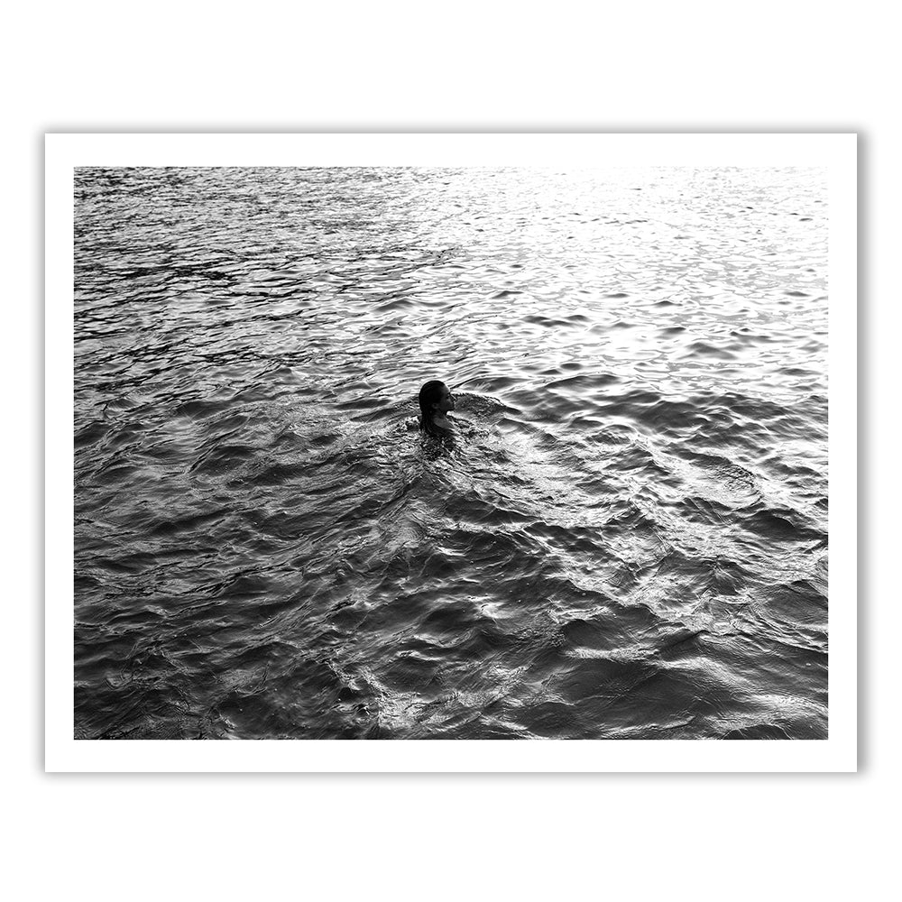 Sunrise Swim #3 by Francesca Owen captures a serene black and white photograph of a swimmer cutting through rippling water with the sun's reflection casting a gentle shimmer on the surface. The swimmer’s head and shoulders are visible as they move gracefully through the tranquil water. Available as fine art prints with worldwide shipping.