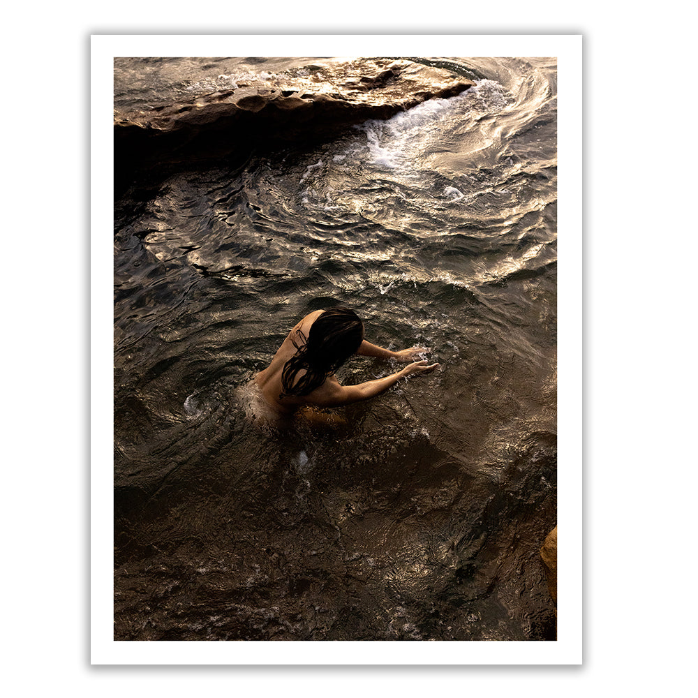 In Francesca Owen's "Sunrise Swim #9," a person with long hair is immersed in swirling dark water, reaching toward a rocky outcrop. The image, taken from above, captures the motion of the water and the texture of the rocks. Printed with archival pigments, this scene's dim lighting gives it a dramatic and mysterious effect.