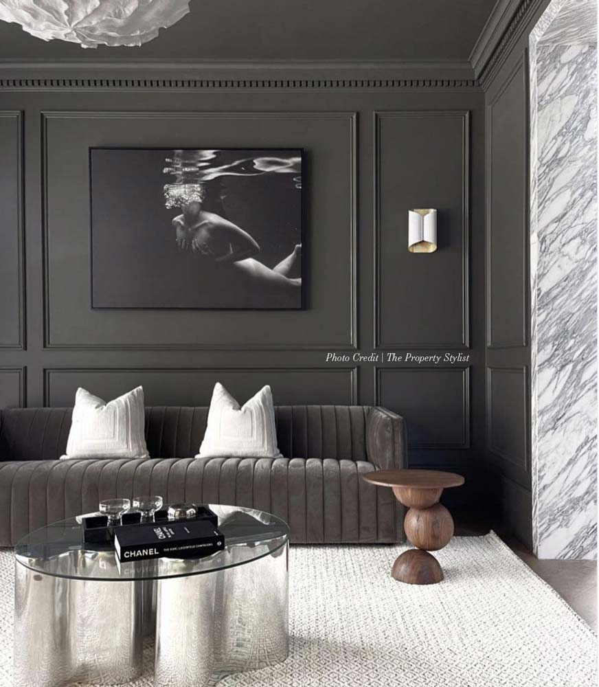 A modern living room features dark gray walls adorned with a large, hand-signed black-and-white photograph titled "Hold On" by Francesca Owen, printed on fine art paper. The room includes a gray tufted couch with white pillows, a round metallic coffee table with books on it, and a wooden side table. A marble accent wall is visible on the right.