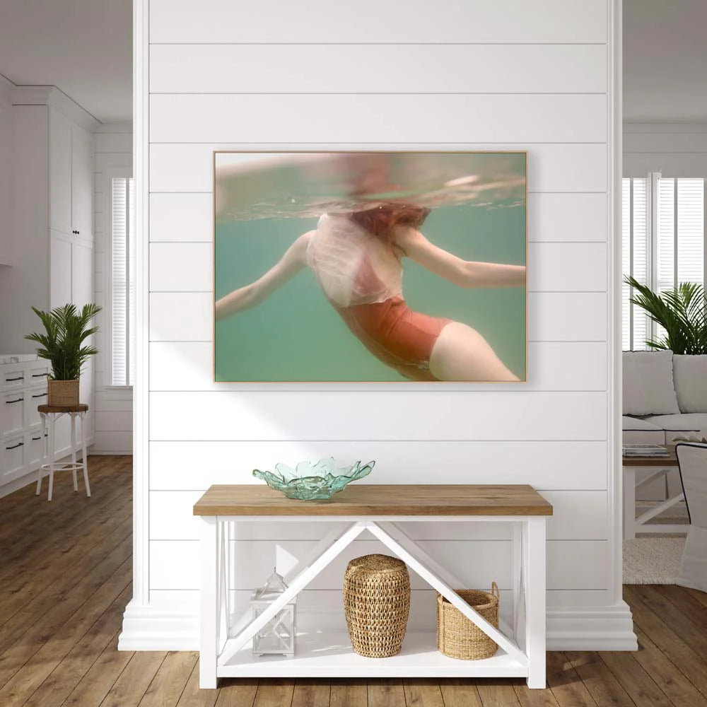 A contemporary living room with white shiplap walls features "Feminine," a large photograph of a person swimming underwater, hand-signed by Francesca Owen for that personal touch. Below the artwork is a wooden console table holding decorative items, while potted plants add a breath of life to the space.