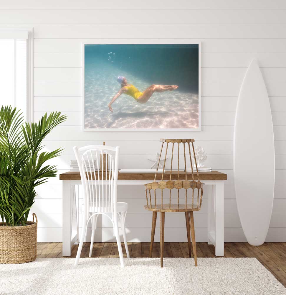 A coastal-themed room with white walls and light wood furniture. The space features a wooden table with white and natural wood chairs, a framed underwater photo titled "Halcyon Daze 2" by Francesca Owen using archival pigments on the wall, a surfboard propped in the corner, and a potted plant on the floor.