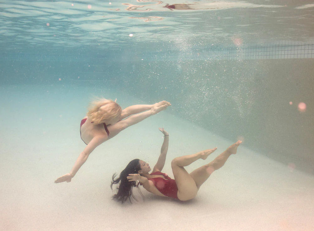 Two women in custom-made red swimsuits from Francesca Owen's "Opposites Attract 02" collection perform a mesmerizing synchronized swimming routine underwater. One is mid-air in a split leap while the other gracefully extends her arms and legs below her. This elegant scene shimmers through the clear water like fine art paper, evoking the signature artistry of Francesca Owen.
