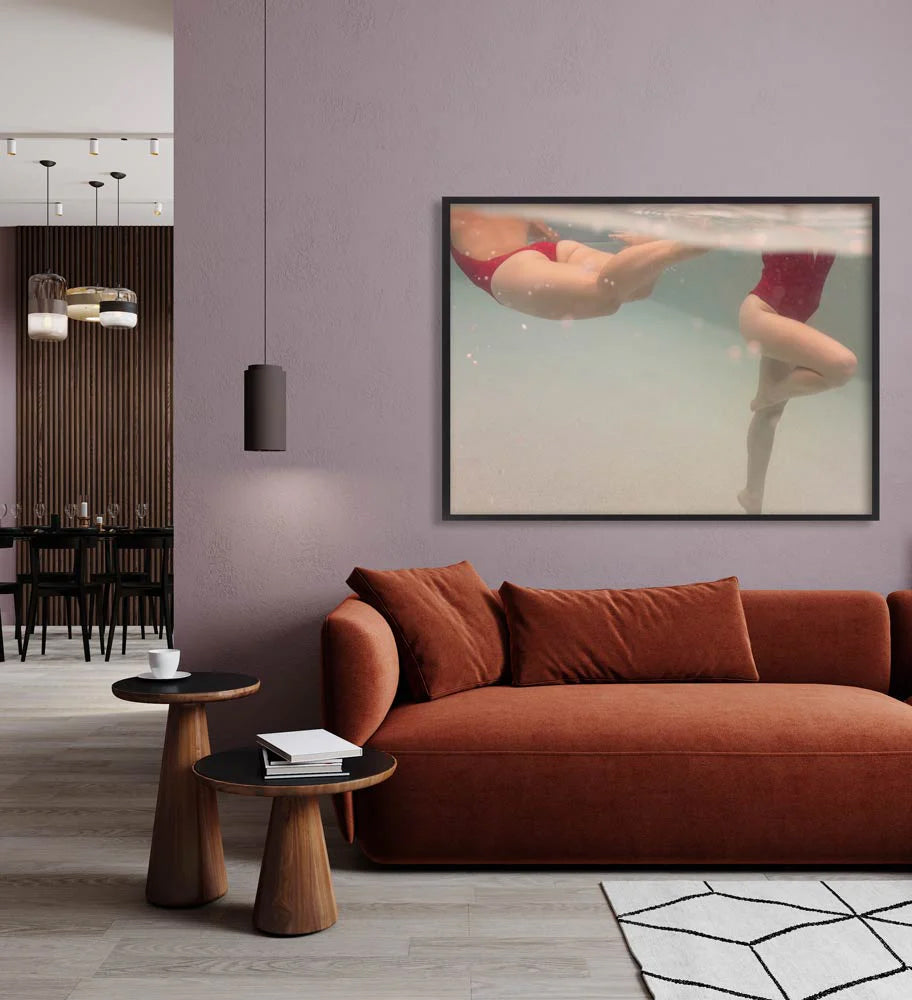 A modern living room showcases a rust-colored sofa and wooden side tables. Prominently displayed on the wall is "Opposites Attract 03," a framed underwater photo of two people swimming, printed on fine art paper by Francesca Owen. Pendant lights hang from the ceiling, and a dining area is visible in the background.