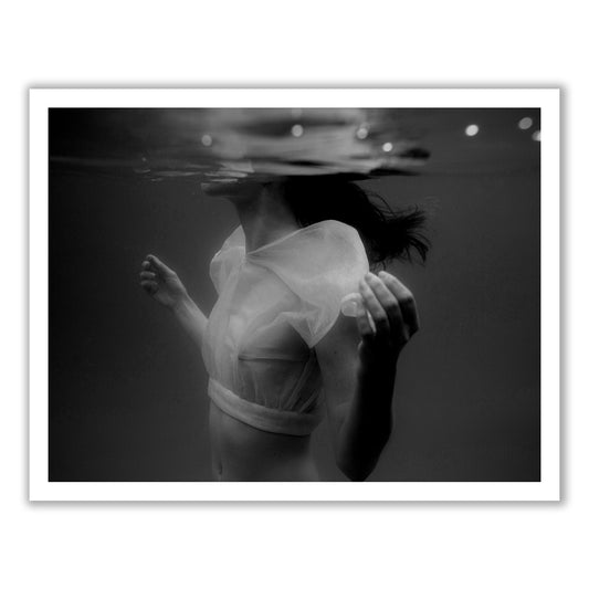 The artwork "Accepting" by Francesca Owen captures a black-and-white photo of a person submerged in water up to their neck, wearing a translucent top with arms gently curved at their sides. Their head is partially above the water's surface, hair floating around. Printed on fine art paper using archival pigments, the mood is ethereal and serene.