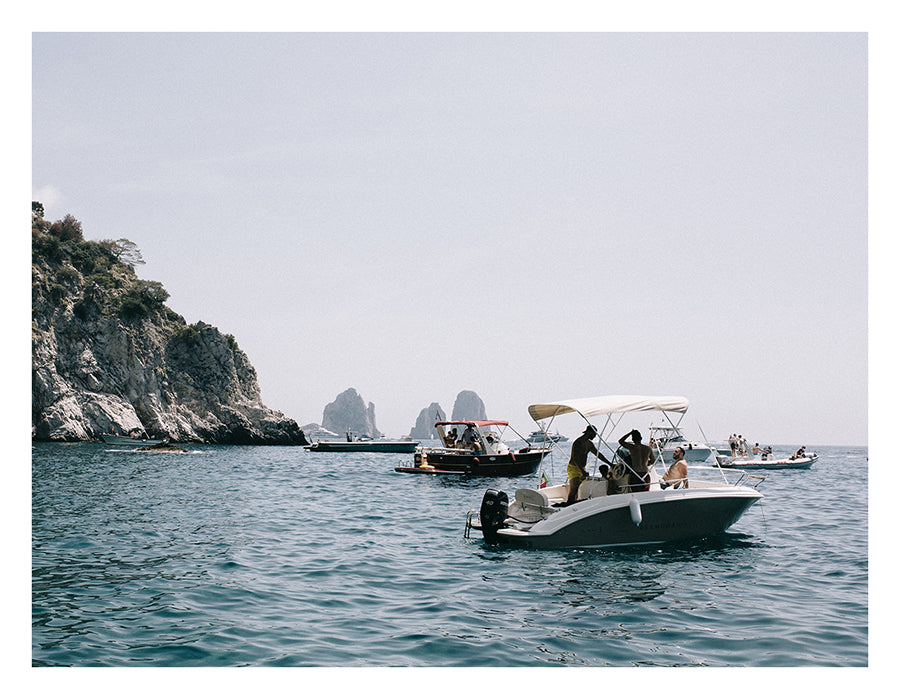 Several small motorboats with people on board are floating on the clear, calm blue water near a rocky coastline in an idyllic scene. In the background, large rock formations rise majestically from the sea under a clear sky, creating a vista reminiscent of "Amalfi By Sea" by Francesca Owen, resembling an image printed on fine art paper using archival pigments.