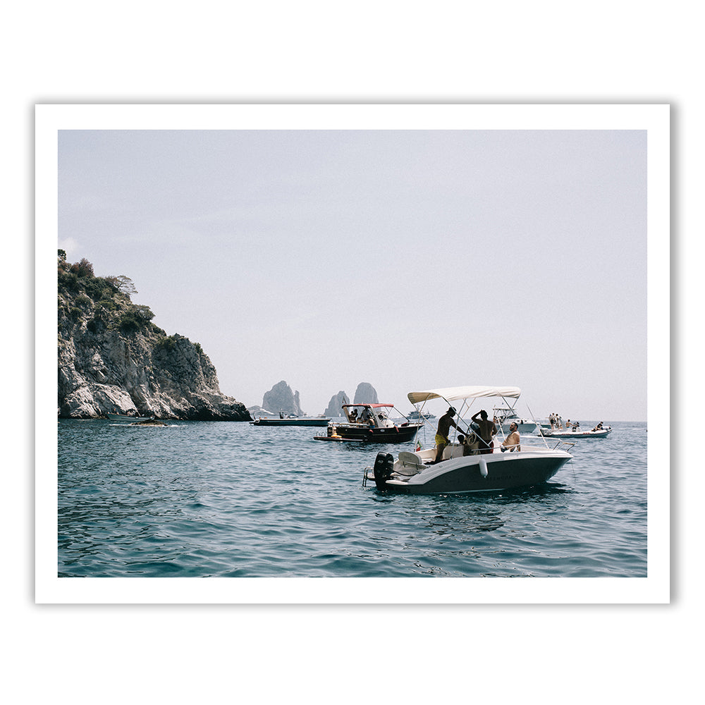 Several small boats float on a calm sea near a rocky coastline. The sky is clear, with distant rock formations visible. In the foreground, some people are seen on a boat, enjoying the serene waters. This beautiful scene is captured in "Amalfi By Sea" by Francesca Owen, printed with archival pigments on hand-signed fine art paper.